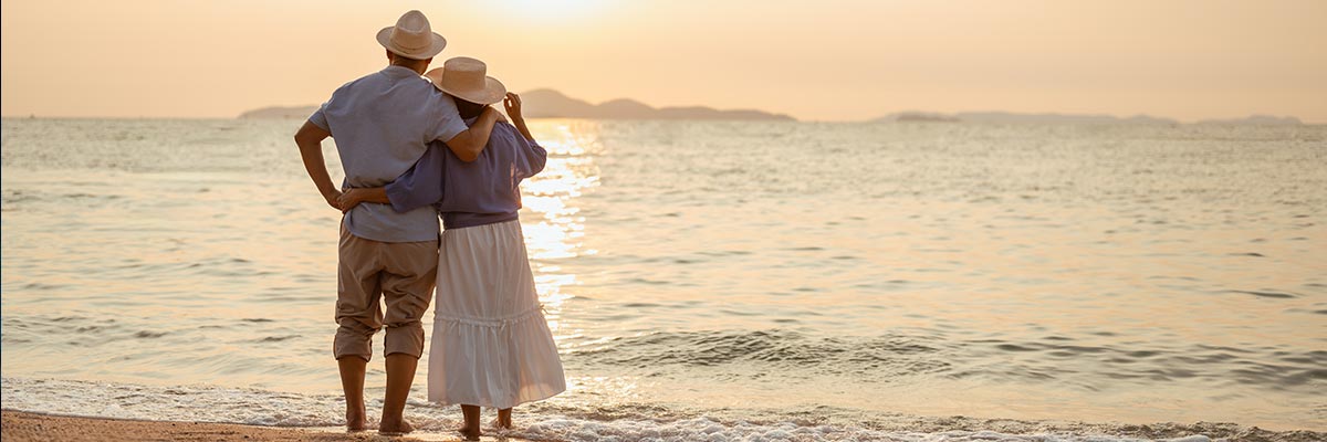 Retired couple standing barefoot at the water's edge in the late afternoon, looking out to sea