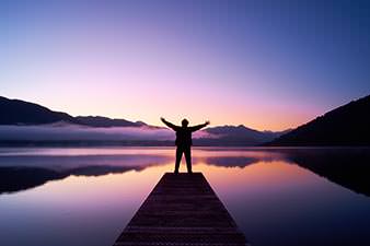 Person standing at the end of a dock looking across Lake Kaniere at sunrise, arms raised to greet the dawn