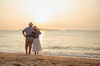 Retired couple standing barefoot at the water's edge in the late afternoon, looking out to sea